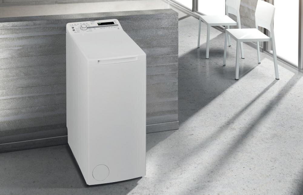 Na obrazie znajduje się biała pralka o pionowym załadunku, ustawiona w nowoczesnym wnętrzu z szarymi ścianami i podłogą. Obok pralki widoczne są dwa białe krzesła przy oknie.