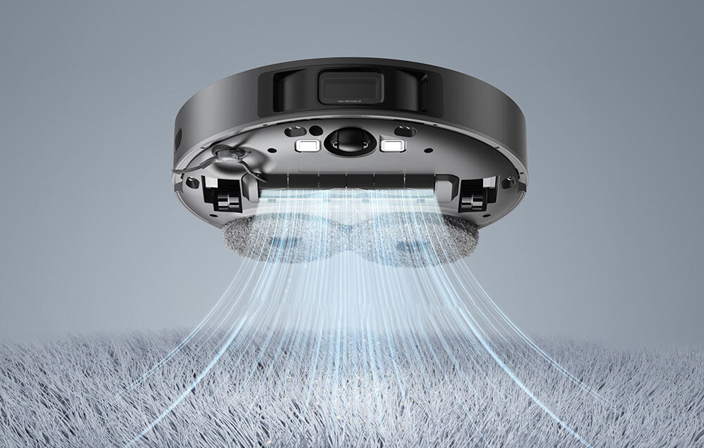 Robot sprzątający unosi się nad powierzchnią, z której wydobywają się niebieskie linie symbolizujące ssanie. Widoczne są elementy konstrukcyjne robota, takie jak szczotki i czujniki.