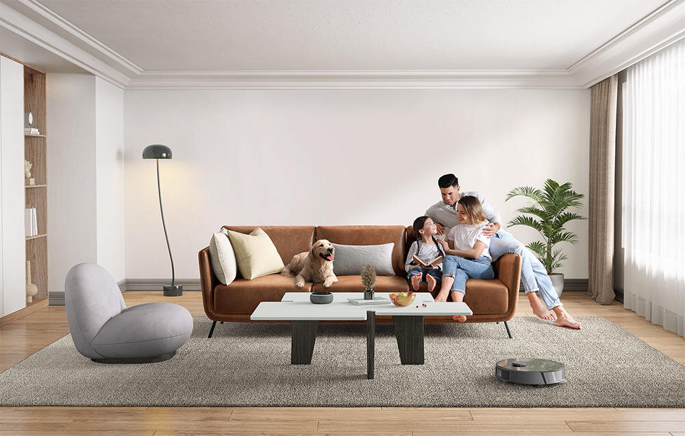 W salonie znajduje się brązowa sofa, na której siedzi rodzina z psem. Na podłodze widoczny jest robot sprzątający, a w tle stoi lampa i roślina doniczkowa.