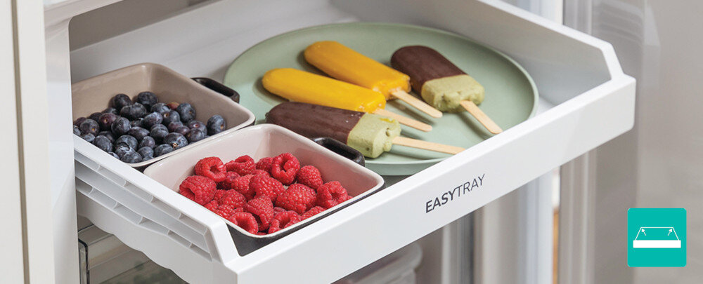Otwarta szuflada lodówki z napisem \'EASYTRAY\', w której znajdują się miseczki z owocami oraz talerz z lodami na patyku. Widać borówki, maliny i lody w różnych kolorach.