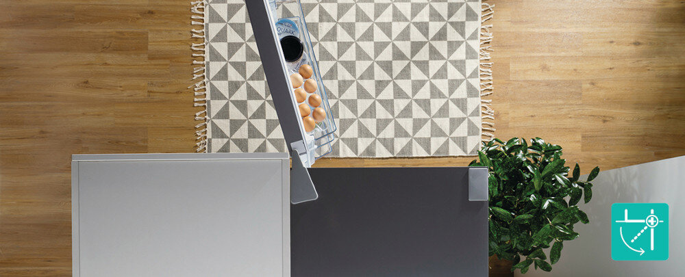 Widok z góry na otwartą szarą lodówkę z widocznymi jajkami i kartonem mleka w środku. Obok lodówki znajduje się roślina doniczkowa i dywan z geometrycznym wzorem na drewnianej podłodze.