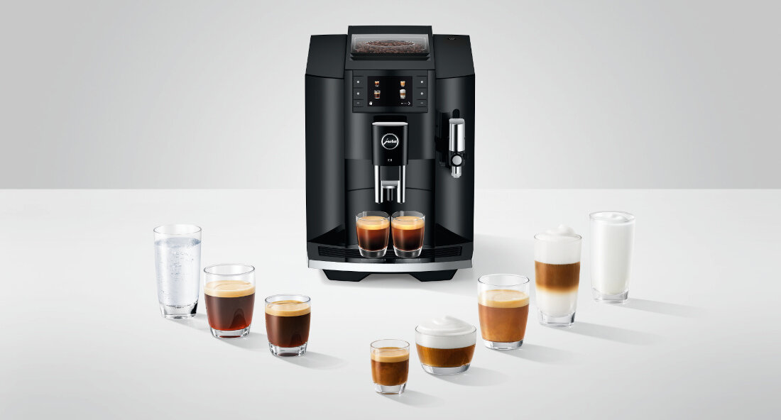 Czarny ekspres do kawy z logo JURA, otoczony różnymi filiżankami z napojami kawowymi i szklanką wody. Ekspres ma wyświetlacz i dyszę do spieniania mleka.