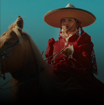 Osoba w czerwonym stroju jeździeckim siedzi na koniu, nosząc duży kapelusz. Tło stanowi jasne, niebieskie niebo.