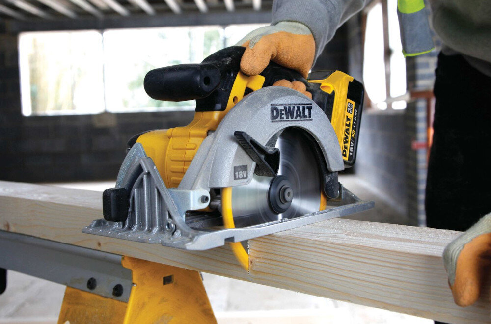 Na zdjęciu widać żółto-szarą pilarkę tarczową marki DEWALT, używaną do cięcia drewnianej belki. Na obudowie narzędzia widoczne są napisy \'DEWALT\' i \'18V\'.