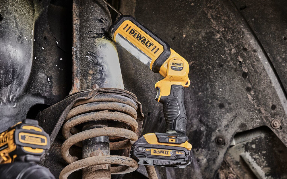 Na obrazie widoczna jest żółto-czarna lampa robocza z logo DEWALT, zamocowana na metalowej konstrukcji. W tle widać elementy mechaniczne pokryte kurzem i brudem.