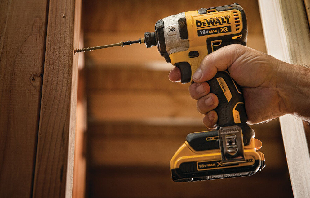 Osoba trzyma w ręku wkrętarkę marki DEWALT, która wkręca śrubę w drewnianą powierzchnię. Na urządzeniu widoczne są napisy: \'DEWALT\', \'18V MAX\', \'XR\'.