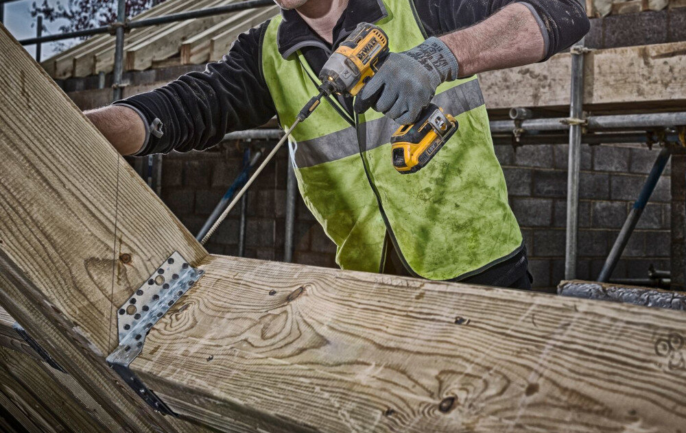 Osoba w kamizelce odblaskowej używa wiertarki akumulatorowej do pracy z drewnianą konstrukcją. W tle widoczne są rusztowania i ceglana ściana.