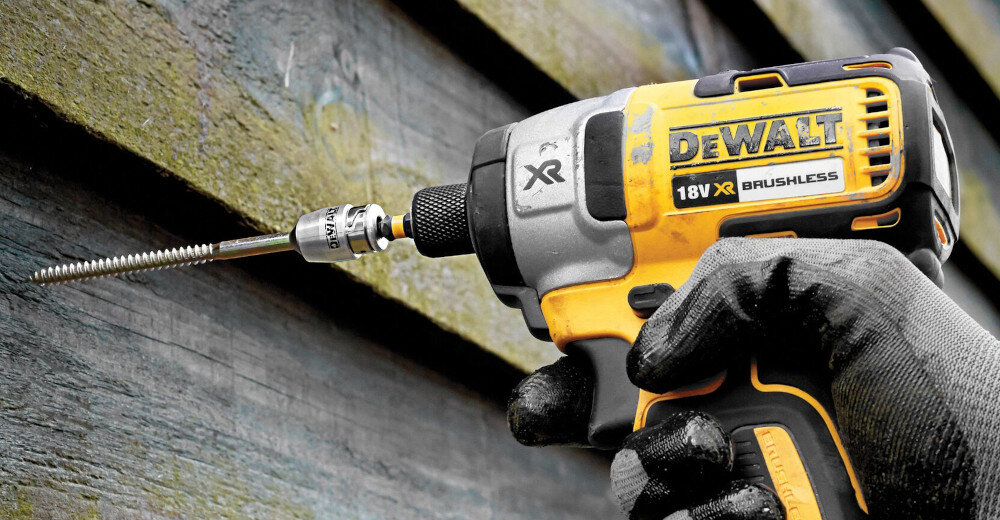 Osoba trzyma w ręku wkrętarkę marki DEWALT z widocznym napisem \'18V XR BRUSHLESS\'. Wkrętarka jest używana do wkręcania śruby w drewnianą powierzchnię.