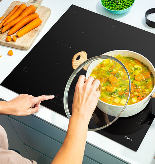 Na obrazie widoczna jest czarna płyta indukcyjna z garnkiem pełnym zupy warzywnej, a obok leżą marchewki na desce do krojenia. Na płycie znajduje się logo \'Amica\'.