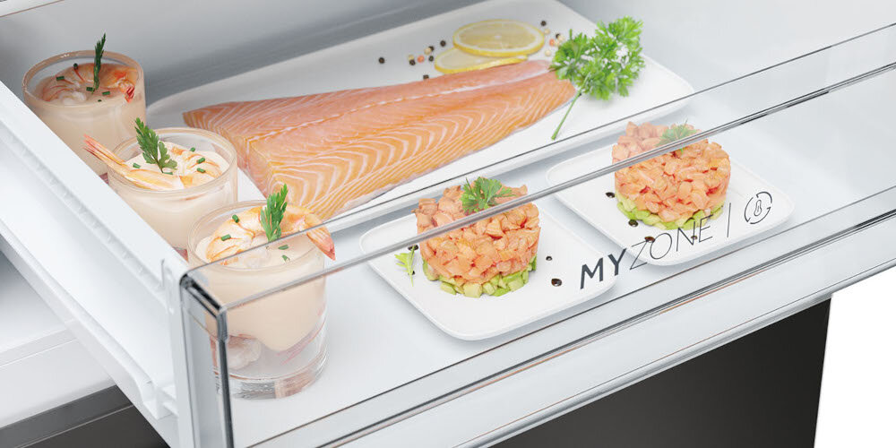 Wnętrze lodówki z półką zawierającą kawałek surowego łososia, plasterki cytryny, gałązkę pietruszki oraz dwa talerzyki z tartarem z łososia. Na szklanej przegrodzie widoczny jest napis \'MY ZONE\'.