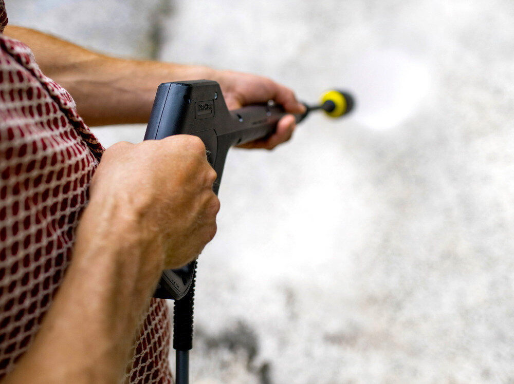 Osoba trzymająca myjkę ciśnieniową, z której wydobywa się strumień wody. Na uchwycie widoczne jest logo \'KARCHER\'.