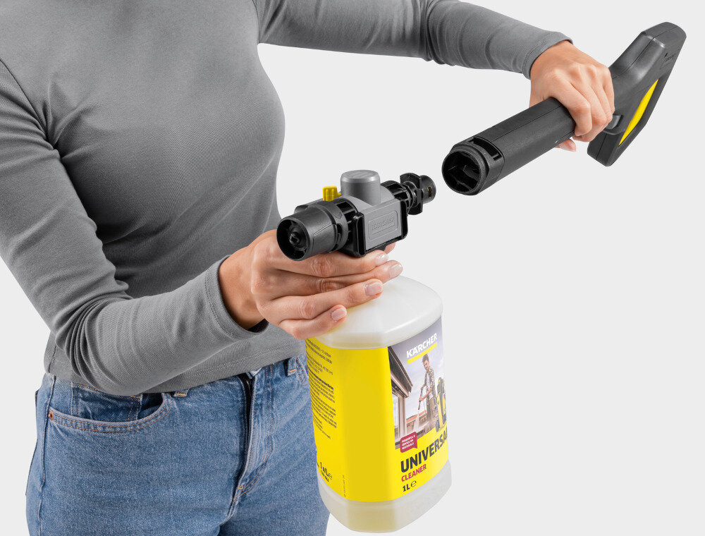 Osoba trzyma myjkę ciśnieniową z podłączonym pojemnikiem na detergent. Na pojemniku widoczny jest napis \'KARCHER UNIVERSAL CLEANER\'.