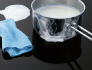 Na płycie indukcyjnej znajduje się garnek z wykipiałym mlekiem, obok leży niebieska ściereczka.