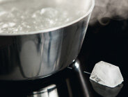 Na obrazie widoczny jest garnek z gotującą się wodą na płycie indukcyjnej oraz kostka lodu obok na powierzchni płyty.