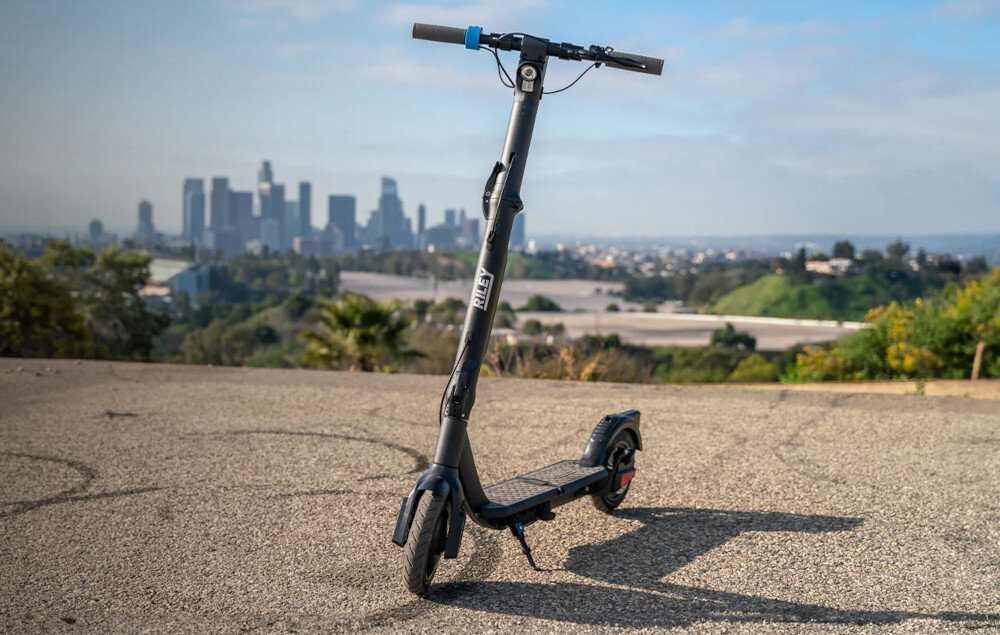 Czarna hulajnoga elektryczna stoi na asfaltowej powierzchni z panoramą miasta w tle. Na kierownicy widoczny jest napis \'RILEY\'.