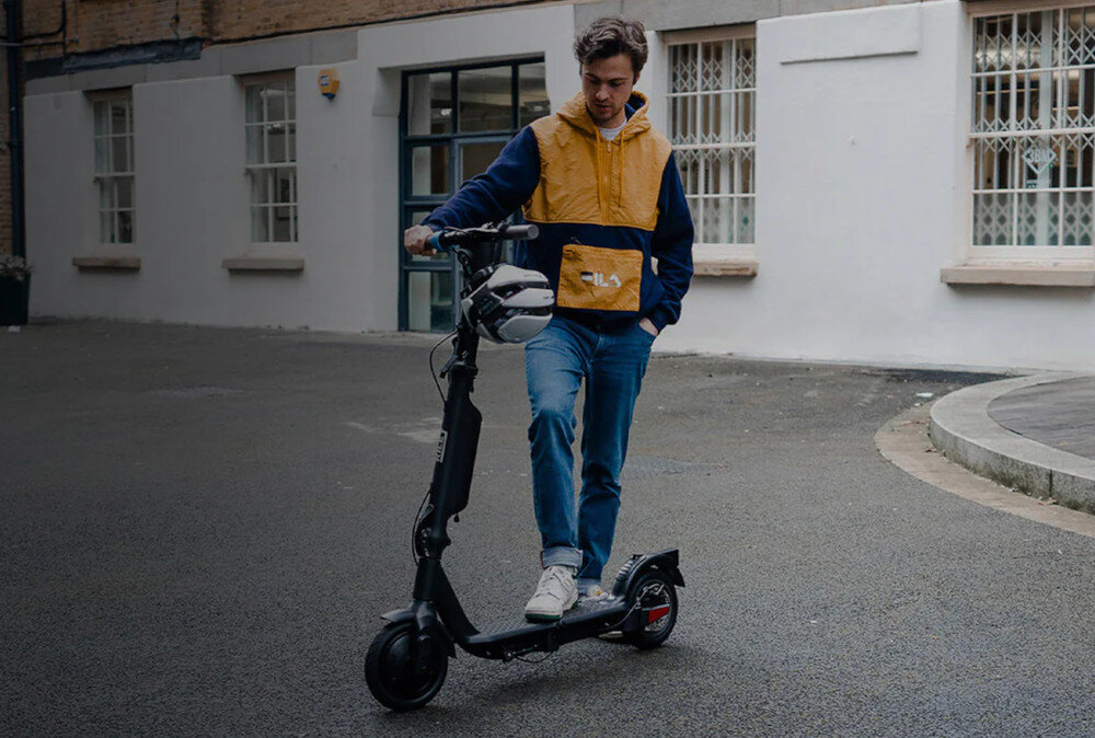 Osoba stoi obok czarnej hulajnogi elektrycznej na tle budynku z oknami. Hulajnoga ma duże koła i kierownicę z zamocowanym kaskiem.