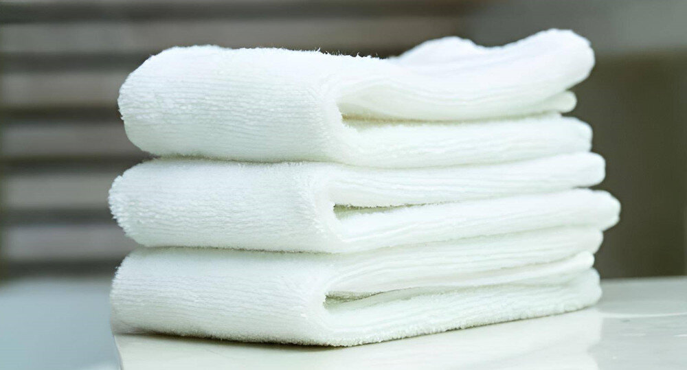 Na obrazie widoczne są trzy białe, złożone ręczniki ułożone jeden na drugim na jasnej powierzchni. W tle znajduje się rozmyte tło w neutralnych kolorach.