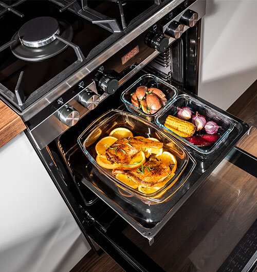 Otwarty piekarnik z dwoma szklanymi naczyniami w środku; w jednym znajduje się pieczone mięso z plasterkami cytryny, w drugim warzywa takie jak kukurydza, cebula i pieczarki. Na panelu sterowania widoczny jest wyświetlacz z cyframi \'14:29\'.