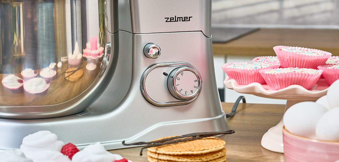 Na blacie kuchennym znajduje się robot planetarny z logo \'zelmer\', obok którego stoją różowe babeczki na paterze oraz wafle i owoce. W tle widać odbicie kuchennych akcesoriów w metalowej powierzchni robota.