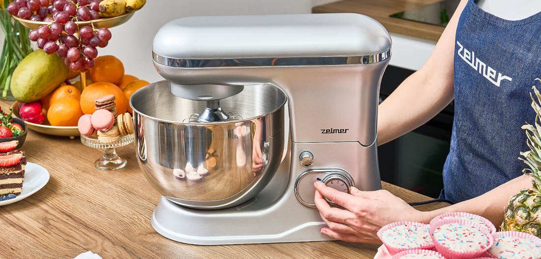 Robot planetarny w kolorze srebrnym z logo \'zelmer\' na obudowie, ustawiony na blacie kuchennym obok owoców i deserów. Osoba w fartuchu obsługuje pokrętło urządzenia.