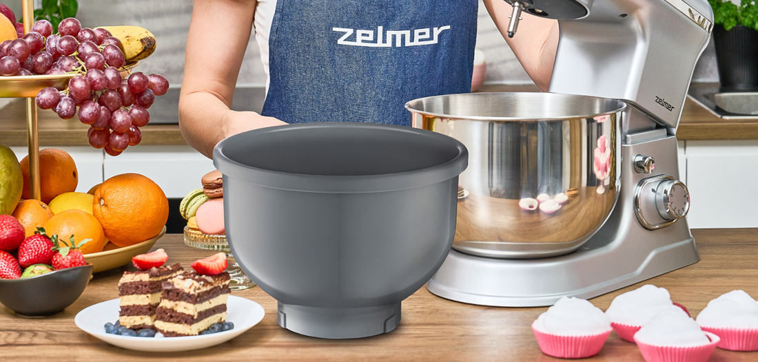 Na blacie kuchennym znajduje się robot planetarny z metalową misą oraz dodatkowa szara misa. W tle widoczne są owoce, ciasto na talerzu i osoba w fartuchu z logo \'zelmer\'.