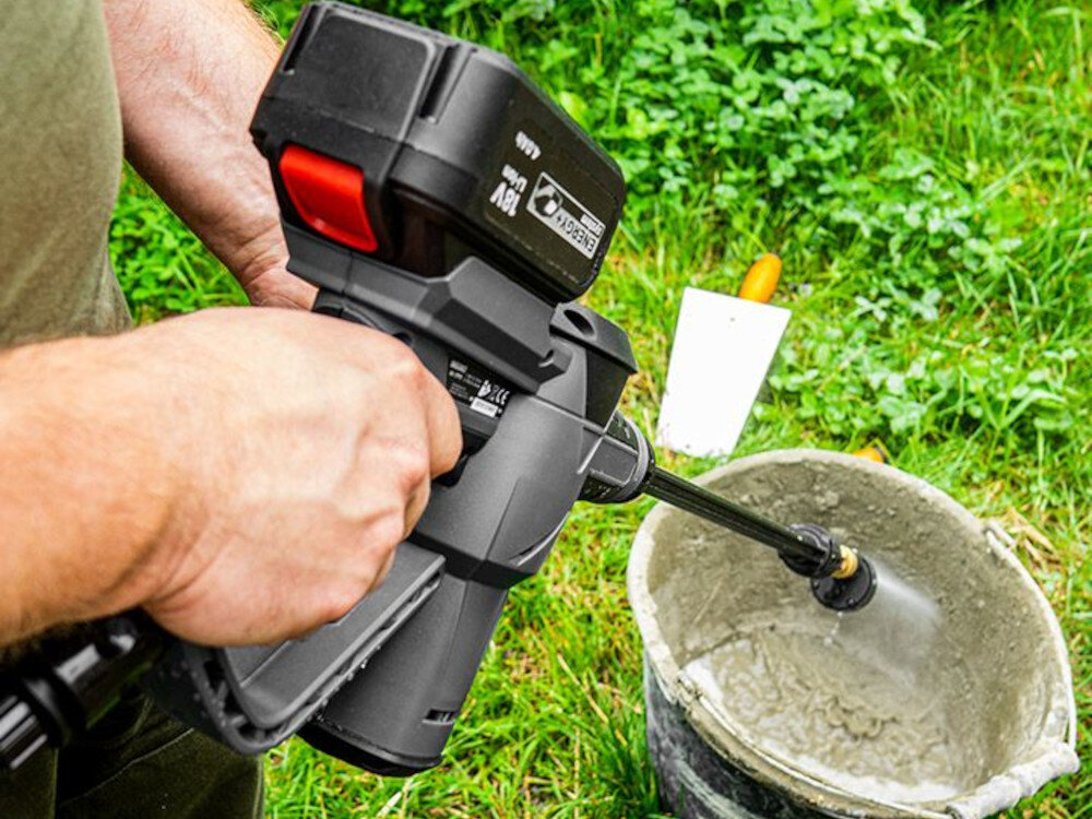 Osoba trzyma myjkę ciśnieniową GRAPHITE Energy+ 58G083, skierowaną na wiadro z wodą, na tle trawy. Na urządzeniu widoczne jest logo \'GRAPHITE 18V\'.