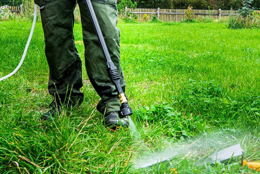 Osoba używa myjki ciśnieniowej do czyszczenia trawnika. W tle widoczny jest drewniany płot i zieleń.