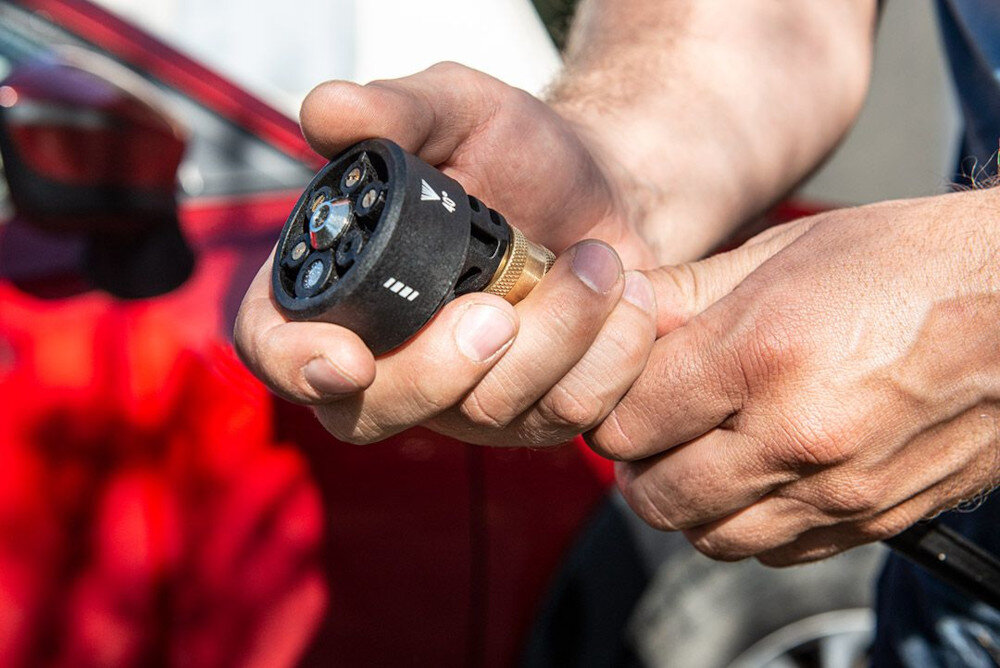 Osoba trzyma w rękach końcówkę myjki ciśnieniowej z widocznymi symbolami i metalowym złączem. W tle znajduje się czerwony samochód.