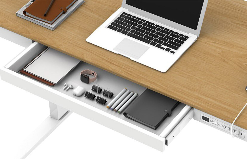 Na biurku z drewnianym blatem znajduje się otwarty laptop oraz notatnik. Pod blatem widoczna jest wysunięta szuflada z różnymi akcesoriami biurowymi, takimi jak długopisy, notes, zegarek i spinacze.