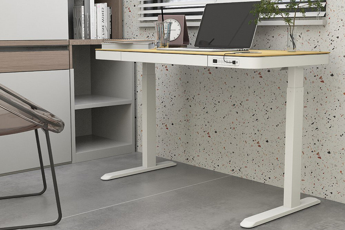 Na obrazie znajduje się biurko w kolorze biało-brązowym z laptopem, szklanką i rośliną na blacie. W tle widać szafki i ścianę z wzorem w kolorowe kropki.