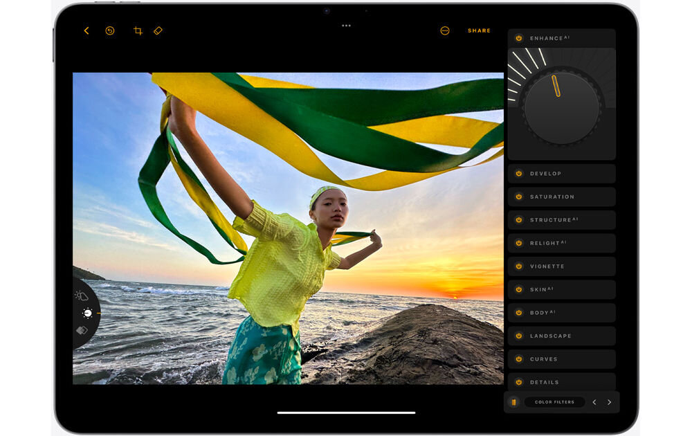 Tablet wyświetla zdjęcie osoby na tle morza i zachodzącego słońca, z zielono-żółtymi wstążkami w rękach. Po prawej stronie ekranu widoczne są opcje edycji zdjęcia, takie jak \'ENHANCE\', \'DEVELOP\', \'SATURATION\'.