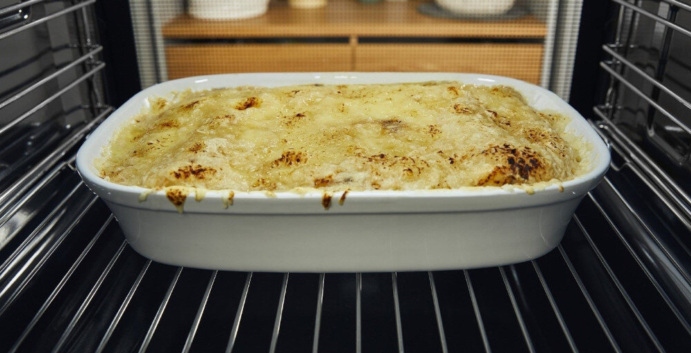 Wnętrze piekarnika z białą ceramiczną formą do pieczenia, w której znajduje się zapiekanka z przypieczonym serem na wierzchu.