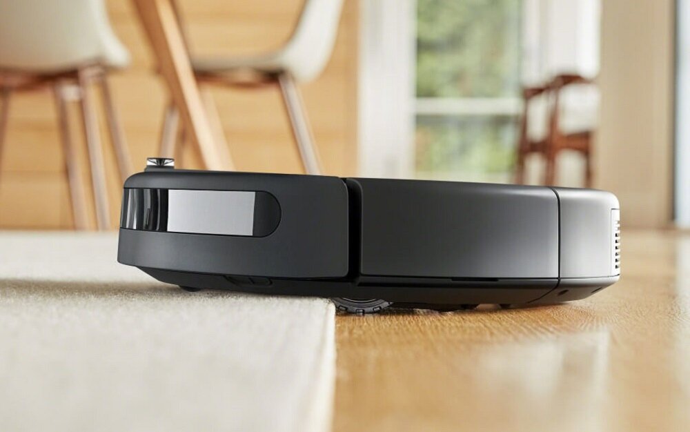 Robot sprzątający w kolorze czarnym pokonuje próg między podłogą a dywanem. W tle widoczne są nogi krzeseł i rozmyte wnętrze pomieszczenia.