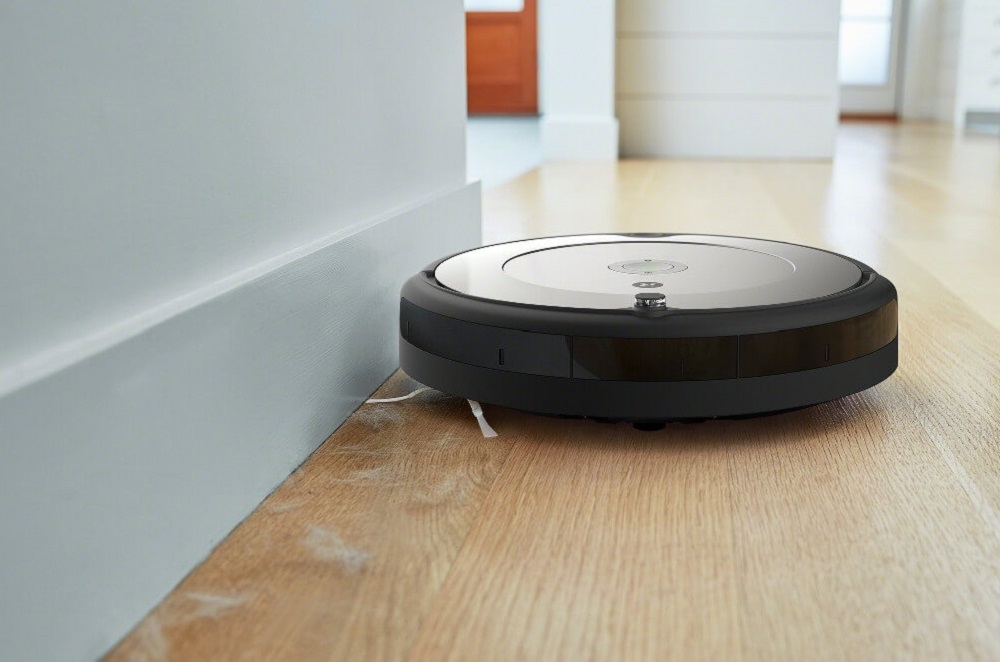 Robot sprzątający porusza się wzdłuż ściany na drewnianej podłodze. Na powierzchni robota widoczne jest logo producenta.