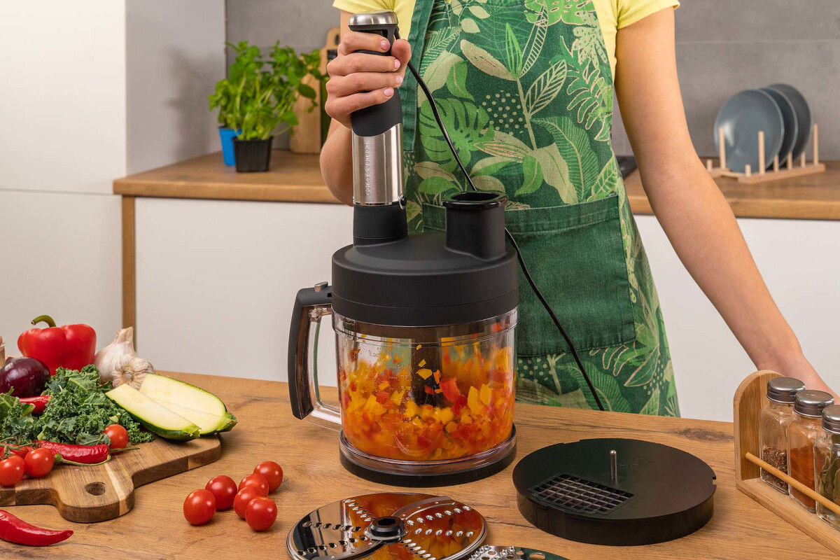 Osoba trzyma blender ręczny z pojemnikiem wypełnionym pokrojonymi warzywami, na blacie znajdują się różne warzywa i akcesoria kuchenne. W tle widać zioła w doniczce i talerze na stojaku.