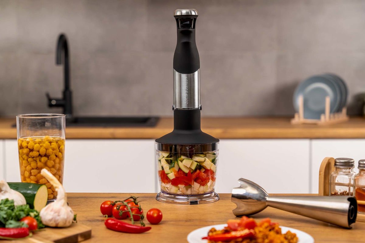 Na blacie kuchennym stoi blender z przezroczystym pojemnikiem wypełnionym pokrojonymi warzywami. W tle widoczne są inne składniki, takie jak ciecierzyca w szklance, pomidory i przyprawy.