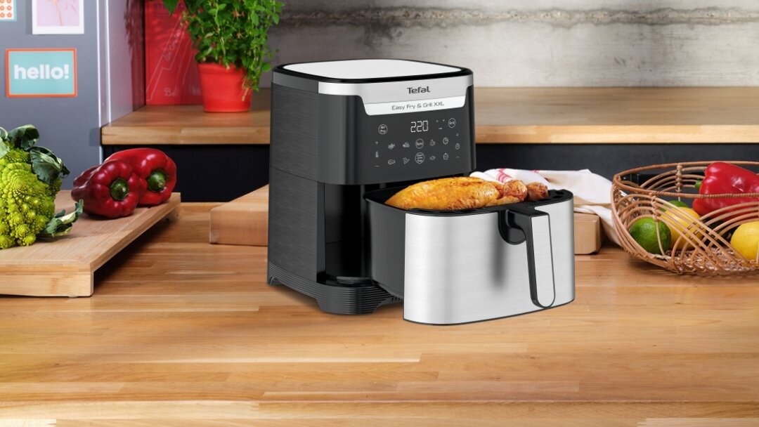 Na blacie kuchennym stoi frytkownica beztłuszczowa TEFAL Easy Fry&Grill XXL EY801D z otwartą szufladą, w której znajduje się pieczony kurczak. W tle widoczne są różne warzywa oraz koszyk z owocami.