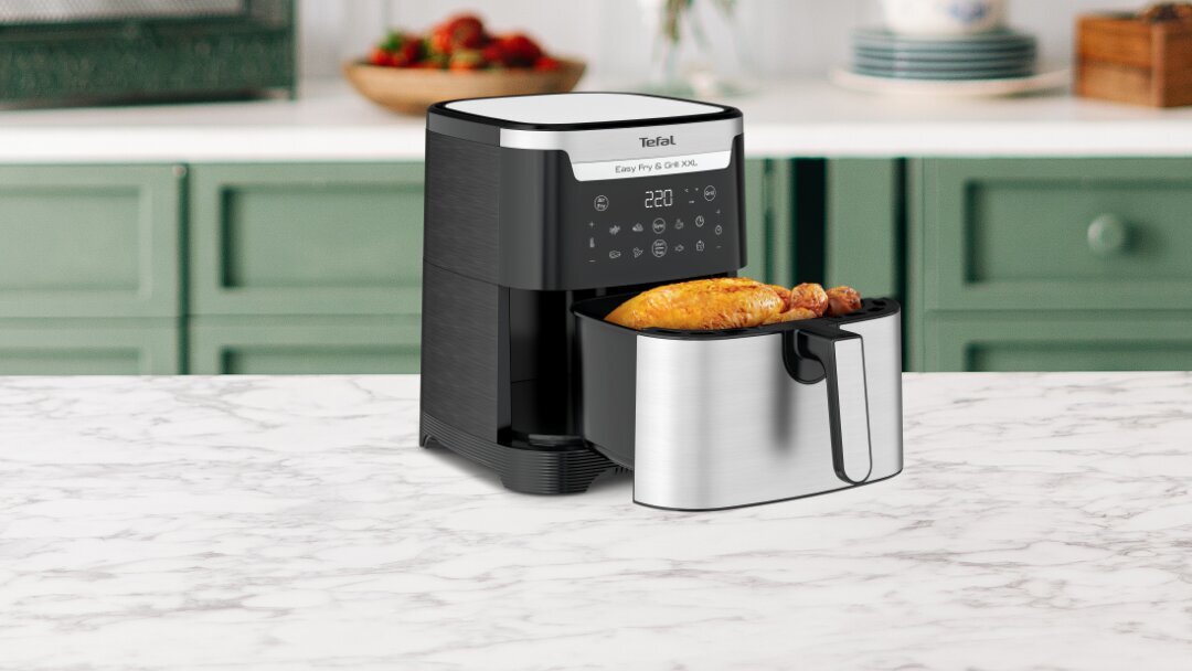 Na blacie kuchennym stoi frytkownica beztłuszczowa z wysuniętą szufladą, w której znajduje się pieczony kurczak. Na panelu urządzenia widoczny jest wyświetlacz z cyfrą 220 i kilkoma ikonami, a na górze napis \'Tefal Easy Fry & Grill XXL\'.