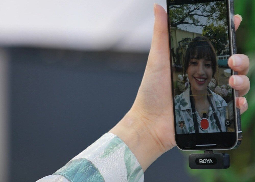 Osoba trzyma smartfon, na ekranie którego widać uśmiechniętą kobietę. Na dole telefonu znajduje się urządzenie z logo \'BOYA\'.