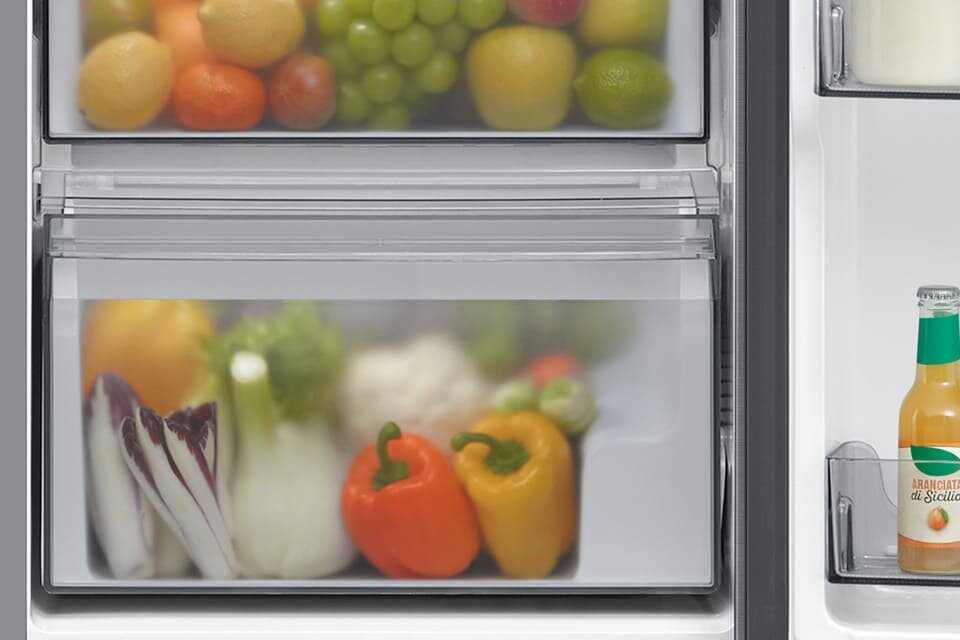 Wnętrze lodówki z widocznymi szufladami wypełnionymi owocami i warzywami, takimi jak papryki i winogrona. Na drzwiach lodówki znajduje się butelka z napisem \'Aranciata di Sicilia\'.