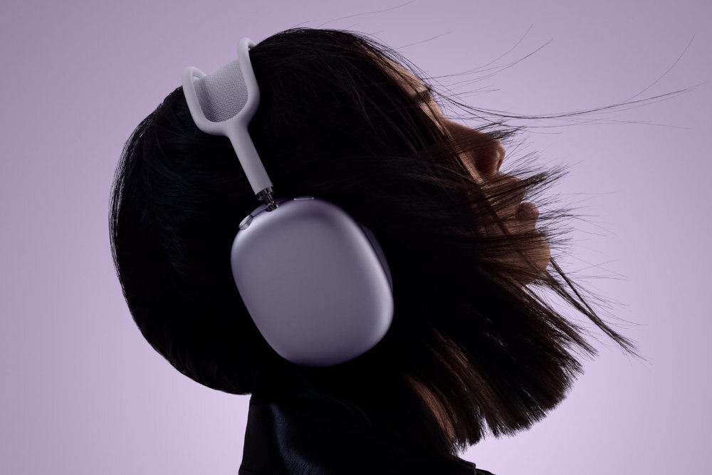 Osoba nosi słuchawki nauszne w kolorze niebieskim na tle fioletowego gradientu. Włosy osoby są rozwiane, a słuchawki mają metaliczny połysk.