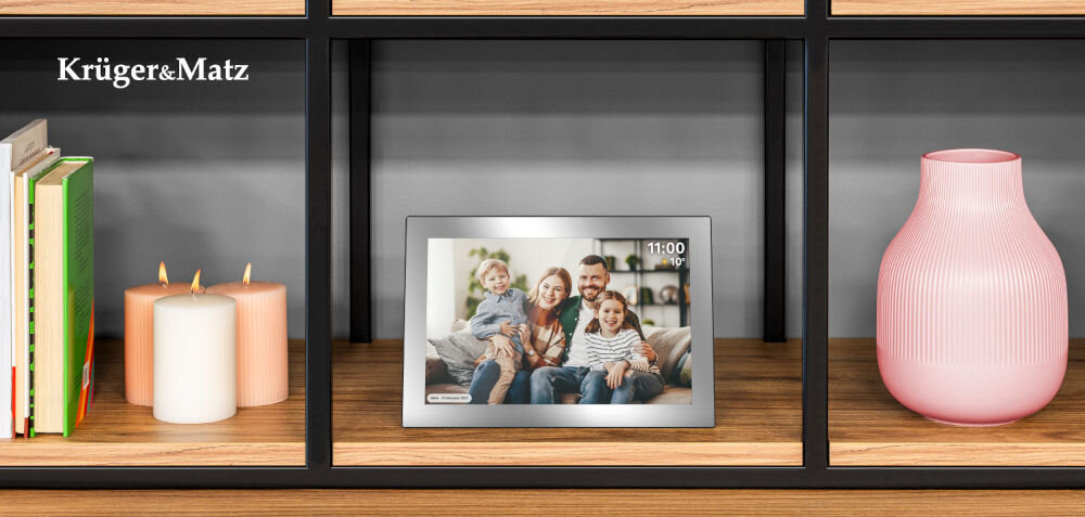 Na półce znajduje się srebrna ramka cyfrowa wyświetlająca zdjęcie rodziny. Obok ramki widoczne są książki, dwie świece i różowy wazon, a w rogu logo Krüger&Matz.