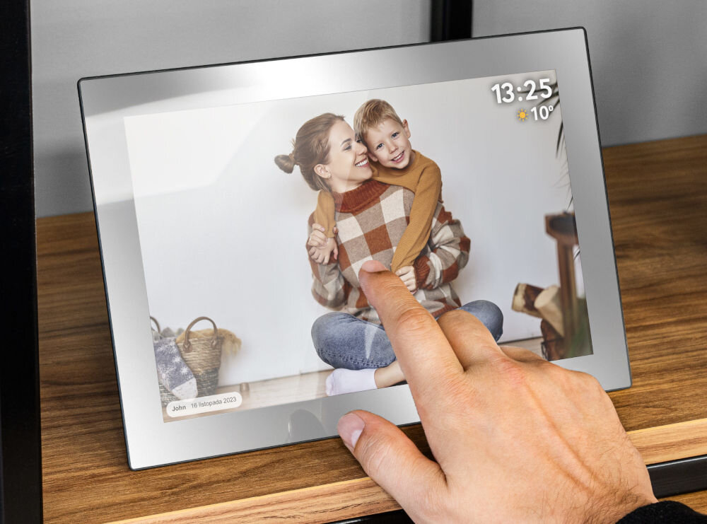 Na zdjęciu widać cyfrową ramkę z wyświetlanym zdjęciem osoby dorosłej i dziecka. W prawym górnym rogu ekranu widoczny jest zegar z godziną 13:25 i temperaturą 10°C, a w lewym dolnym rogu napis \'John 16 listopada 2023\'.