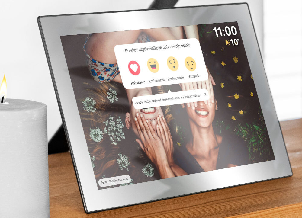 Na zdjęciu widać srebrną ramkę cyfrową z wyświetlaczem, na którym pokazane jest zdjęcie dwóch uśmiechniętych osób oraz interfejs z reakcjami: \'Polubienie\', \'Rozbawienie\', \'Zaskoczenie\', \'Smutek\'. W prawym górnym rogu ekranu widnieje godzina 11:00 i temperatura 10°C.
