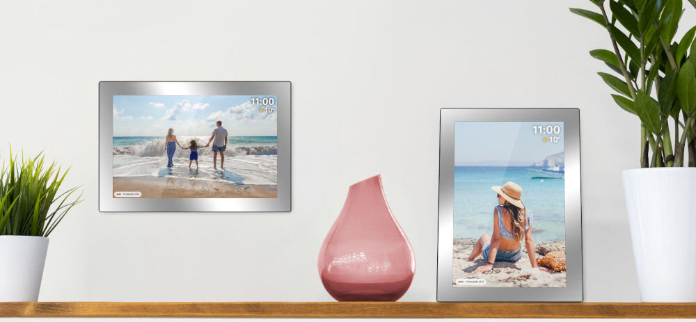 Na półce znajdują się dwie srebrne ramki cyfrowe wyświetlające zdjęcia plażowe, obok różowego wazonu i zielonej rośliny. Na ekranach ramek widoczna jest godzina 11:00 i temperatura 10°C.