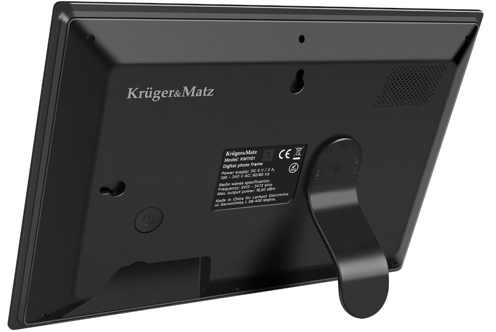 Tył czarnej ramki cyfrowej z logo Krüger&Matz, widoczny uchwyt do postawienia oraz etykieta z informacjami technicznymi. Na etykiecie znajduje się tekst: \'Krüger&Matz Model: KM1101 Digital photo frame Power supply: DC 5V 2A\'.