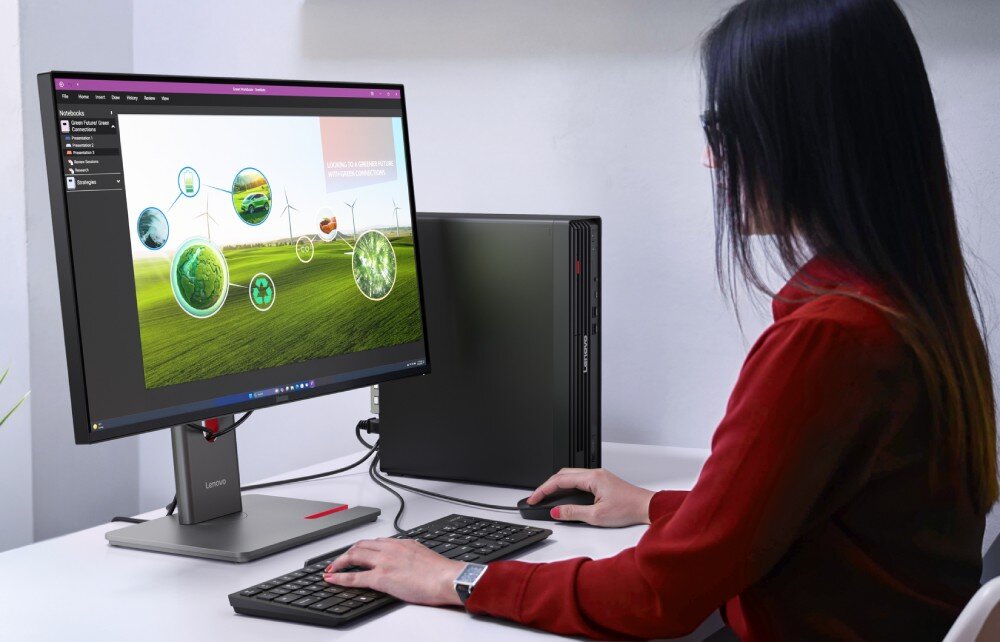 Komputer LENOVO ThinkCentre M90s Gen 6 Kobieta siedzi przy biurku, pisze na klawiaturze i patrzy na monitor stacjonarnego komputera, obok stoi pionowa obudowa