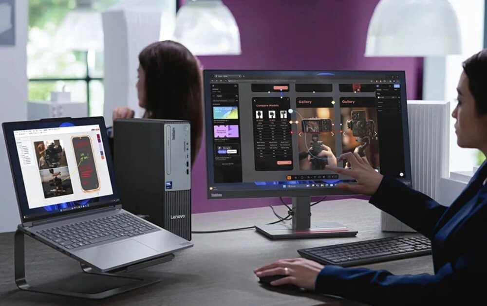 Na biurku znajduje się komputer stacjonarny Lenovo z monitorem i laptopem, na których wyświetlane są różne aplikacje. W tle widoczne są osoby pracujące przy biurkach.