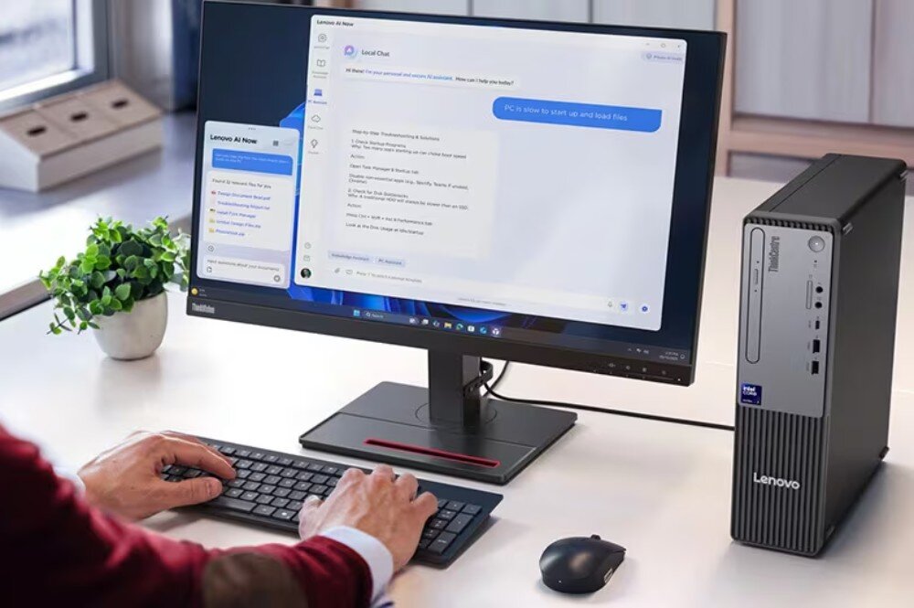 Na biurku znajduje się komputer stacjonarny Lenovo z monitorem, klawiaturą i myszką. Na ekranie widoczny jest interfejs czatu z tekstem: \'PC is slow to start up and load files\'.