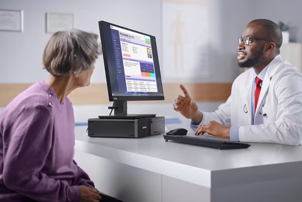Na biurku stoi komputer z monitorem, na którym wyświetlana jest strona internetowa. Obok monitora znajduje się klawiatura, a na obudowie komputera widoczne jest logo Lenovo.